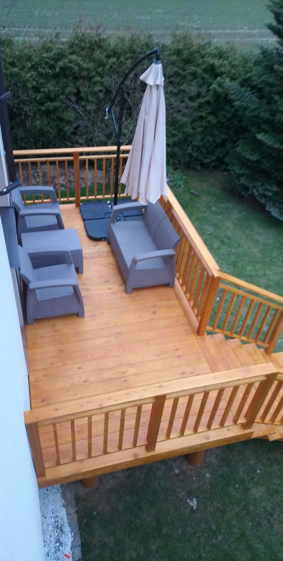 Drewniany taras z meblami ogrodowymi i parasolem, widok z góry. Jasne drewno, szare meble, zielone krzewy w tle. Schody prowadzą na trawnik.