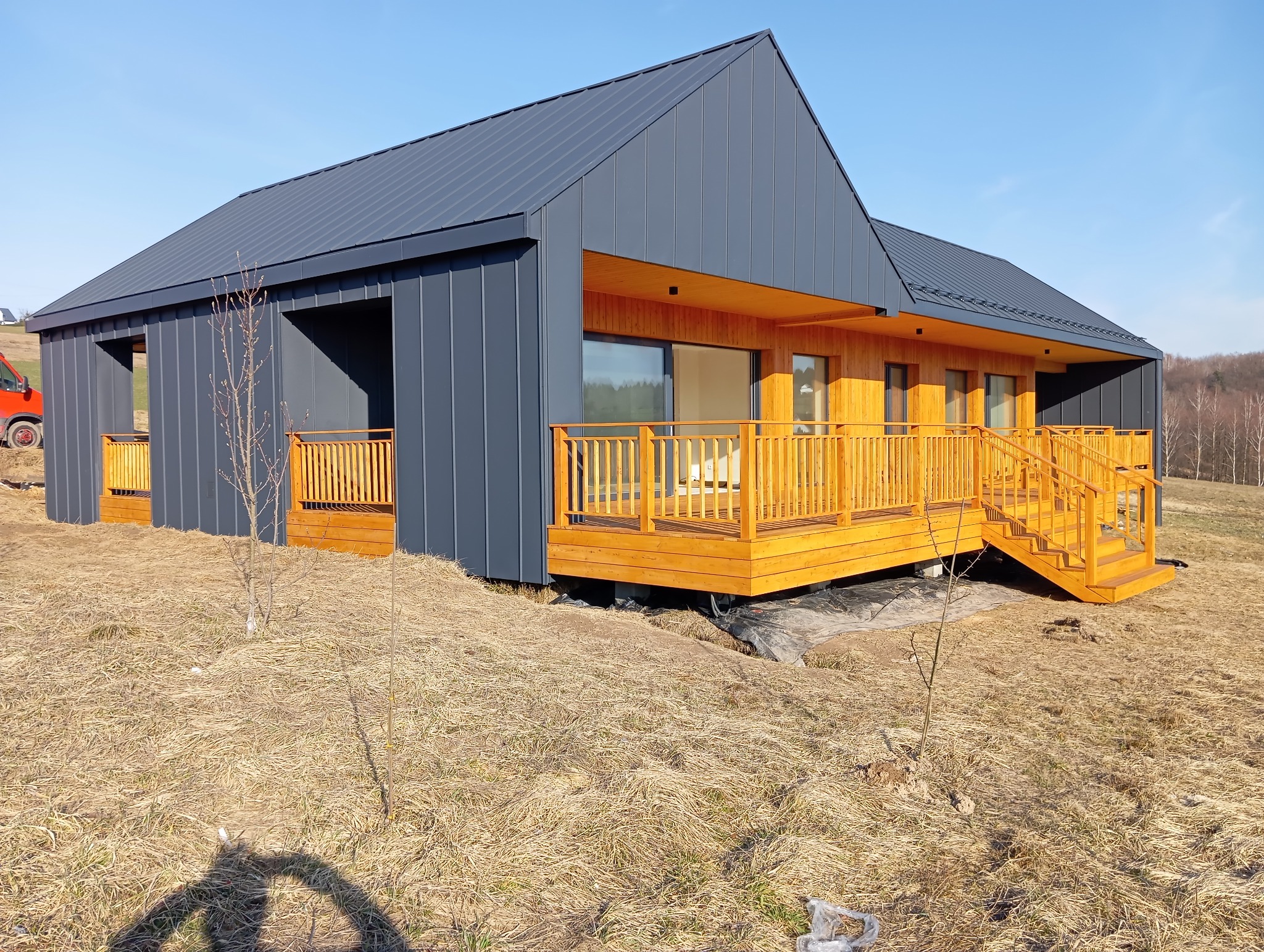 Nowoczesny dom z drewnianym tarasem i szarą elewacją na tle błękitnego nieba. Widoczne schody tarasowe i elementy wykończeniowe.