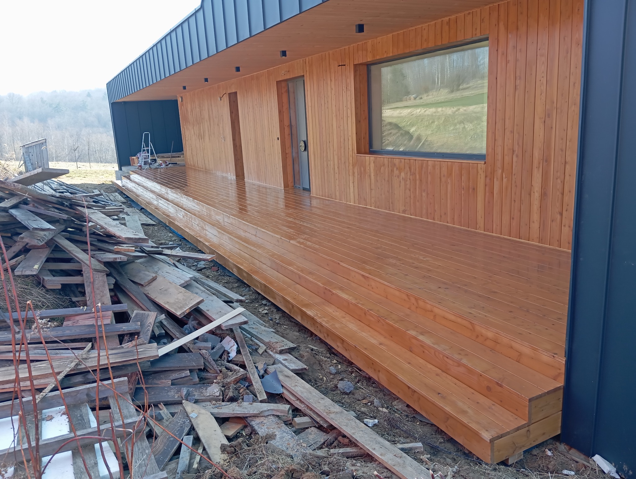 Nowoczesny taras drewniany z widocznymi schodami i fragmentem elewacji budynku. Świeżo zaimpregnowane deski tarasowe. W tle widoczne drzewa i niebo.