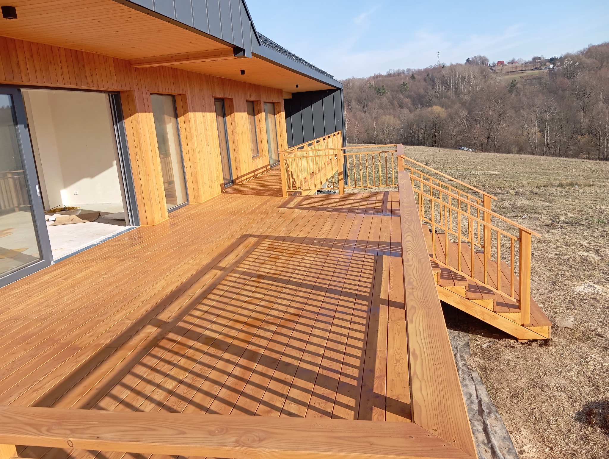 Nowoczesny taras drewniany z balustradą i schodami, przylegający do budynku z drewnianą elewacją i dużymi oknami. Widok na wzgórze w tle.