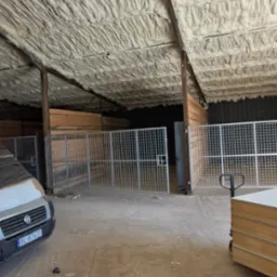 Poddasze budynku gospodarczego po ociepleniu pianką PUR, widoczne metalowe kojce i auto dostawcze w tle. Izolacja natryskowa na drewnianej konstrukcji dachu.