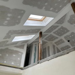 PUROG ADAM ROGOWSKI - Skośny sufit z płyt karton-gips z widocznymi szpachlowaniami, dwa świetliki z drewnianymi ramami i drewniana belka konstrukcyjna w trakcie wykończenia poddasza.