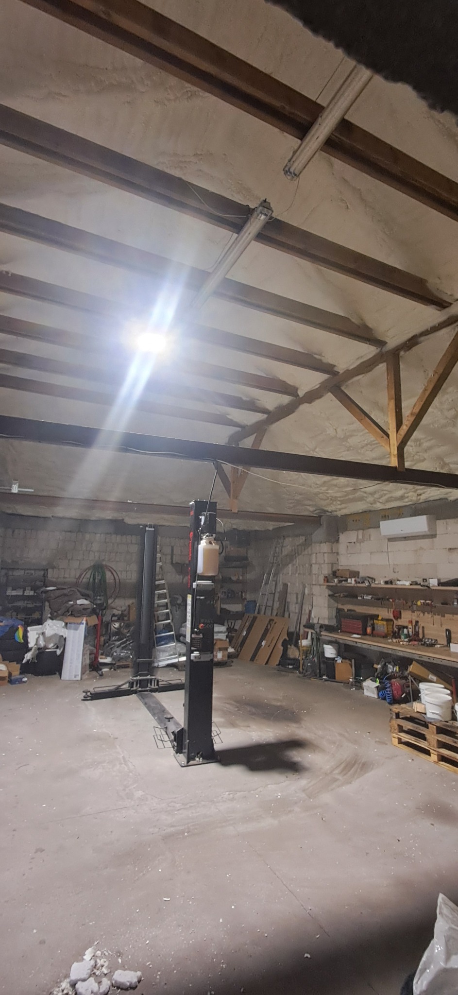 Wnętrze warsztatu z podnośnikiem samochodowym i ocieplonym pianką PUR sufitem z drewnianą konstrukcją. Widoczne narzędzia i materiały warsztatowe.