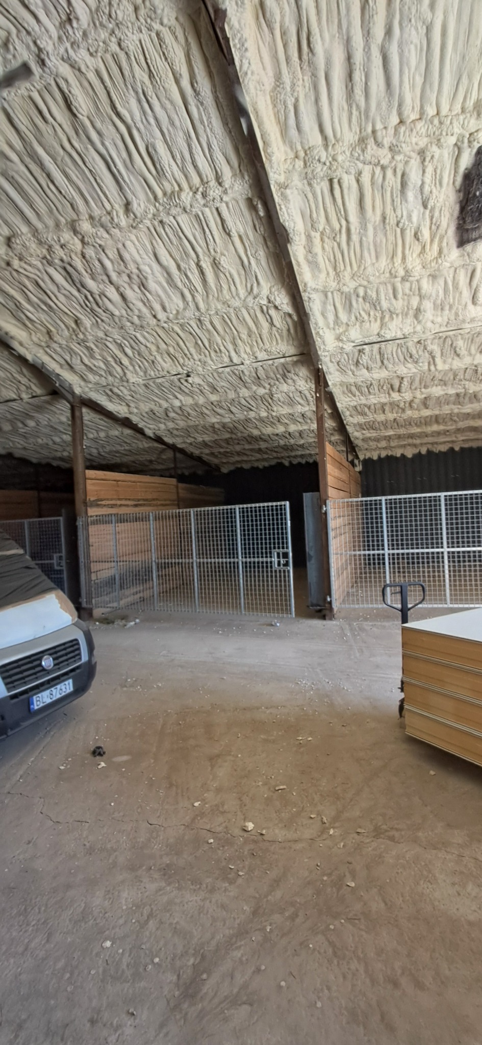 Poddasze budynku gospodarczego po ociepleniu pianką PUR, widoczne metalowe kojce i auto dostawcze w tle. Izolacja natryskowa na drewnianej konstrukcji dachu.