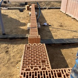 SolidHouse - Wznoszenie fundamentów z pustaków ceramicznych na piasku, widoczne zbrojenie i izolacja. W tle dom w budowie i zaparkowany samochód.