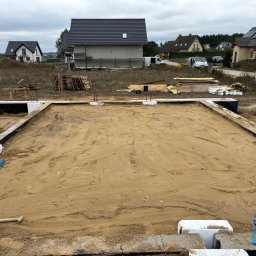 SolidHouse - Przygotowany teren pod fundamenty domu jednorodzinnego. Widoczne zbrojenia, izolacja i piasek. W tle zabudowa jednorodzinna i pochmurne niebo.