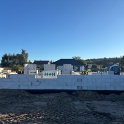 SolidHouse - Wznoszone ściany z betonu komórkowego H+H na fundamencie. Widoczny fragment budynku w trakcie budowy, zarys murów na tle błękitnego nieba i zabudowy jednorodzinnej.