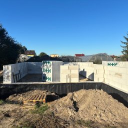 SolidHouse - Wznoszenie ścian fundamentowych z bloczków H+H na czarnej izolacji. Widoczne sterczące pręty zbrojeniowe, paleta i pryzma ziemi. W tle zabudowa jednorodzinna i szklarnia.
