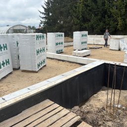 SolidHouse - Fundamenty domu w trakcie budowy: izolacja pionowa, bloczki H+H, zbrojenie. W tle pracownik budowlany i szklarnia. Ujęcie z bliska, widoczne detale izolacji.