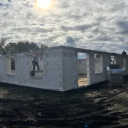 Wznoszenie ścian parterowego domu z betonu komórkowego. Dwóch pracowników na budowie, palety z bloczkami. Widok na fundamenty i częściowo postawione ściany.
