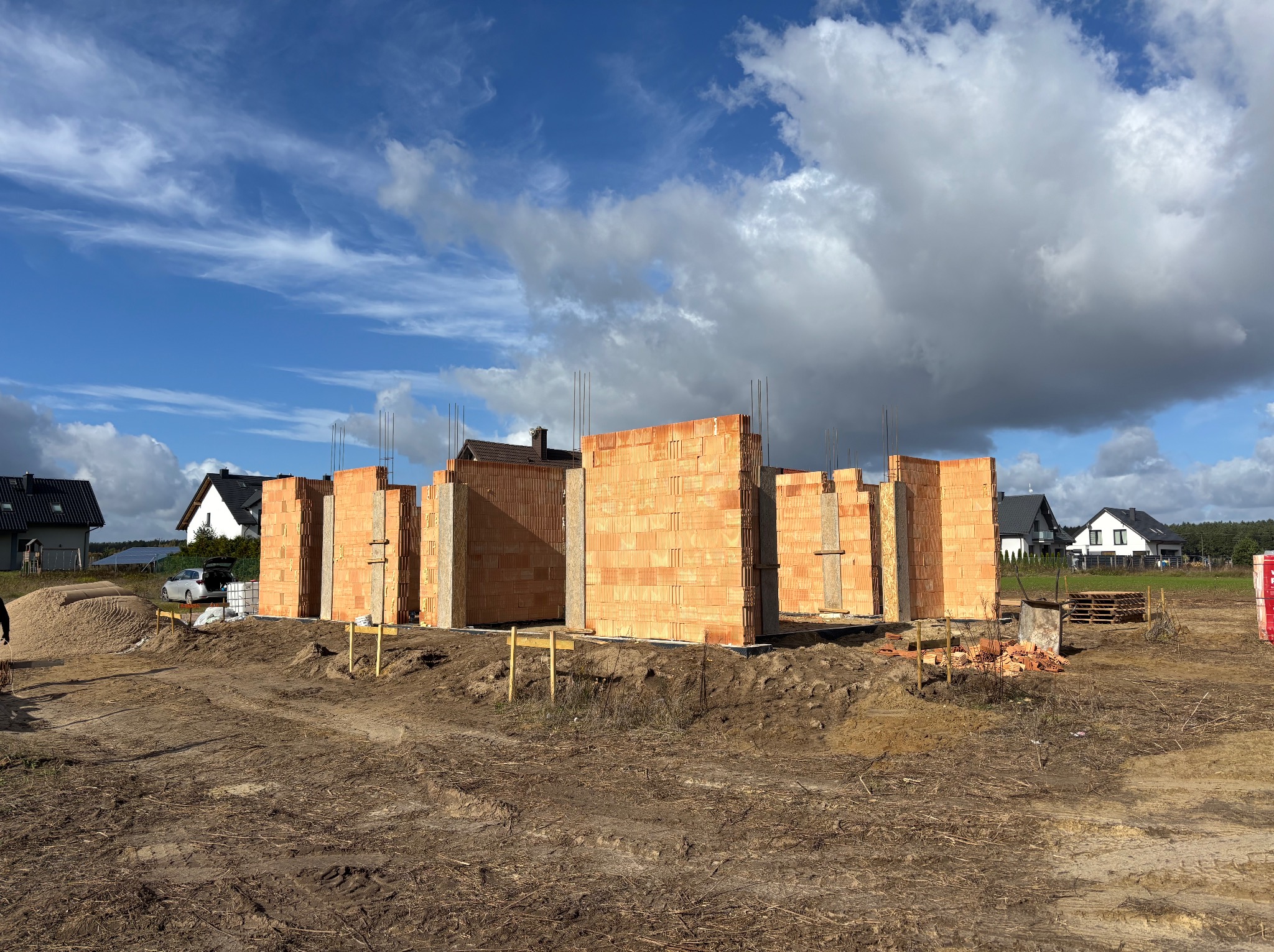 Wznoszone ściany z czerwonej cegły Porotherm na budowie domu jednorodzinnego w słoneczny dzień. Widoczne zbrojenia i drewniane podpory. W tle domy i błękitne niebo.