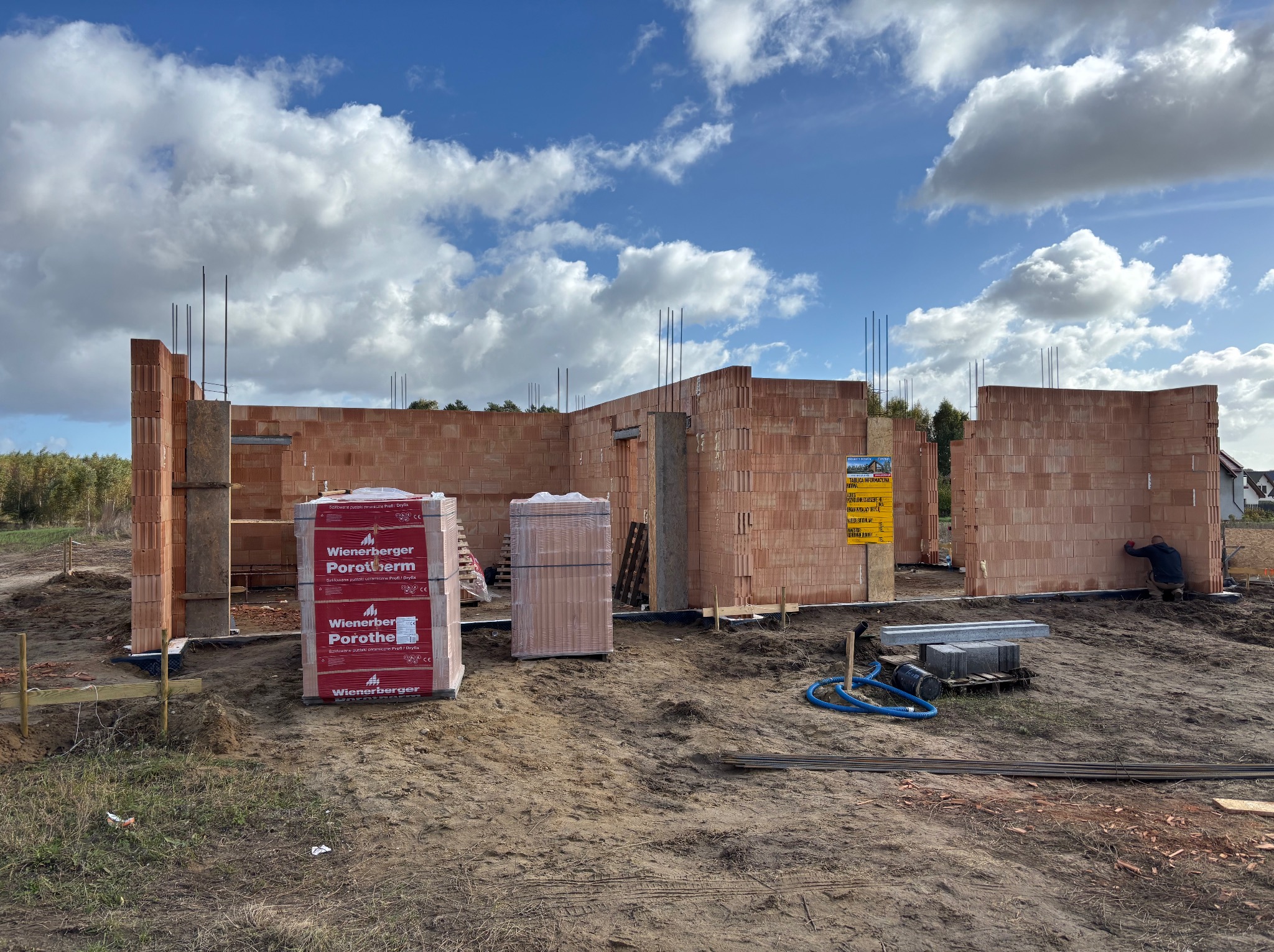 Budowa domu w trakcie: wzniesione ściany z czerwonej cegły Porotherm, widoczne palety z materiałem, narzędzia i pracownik przy murowaniu. Błękitne niebo z chmurami w tle.