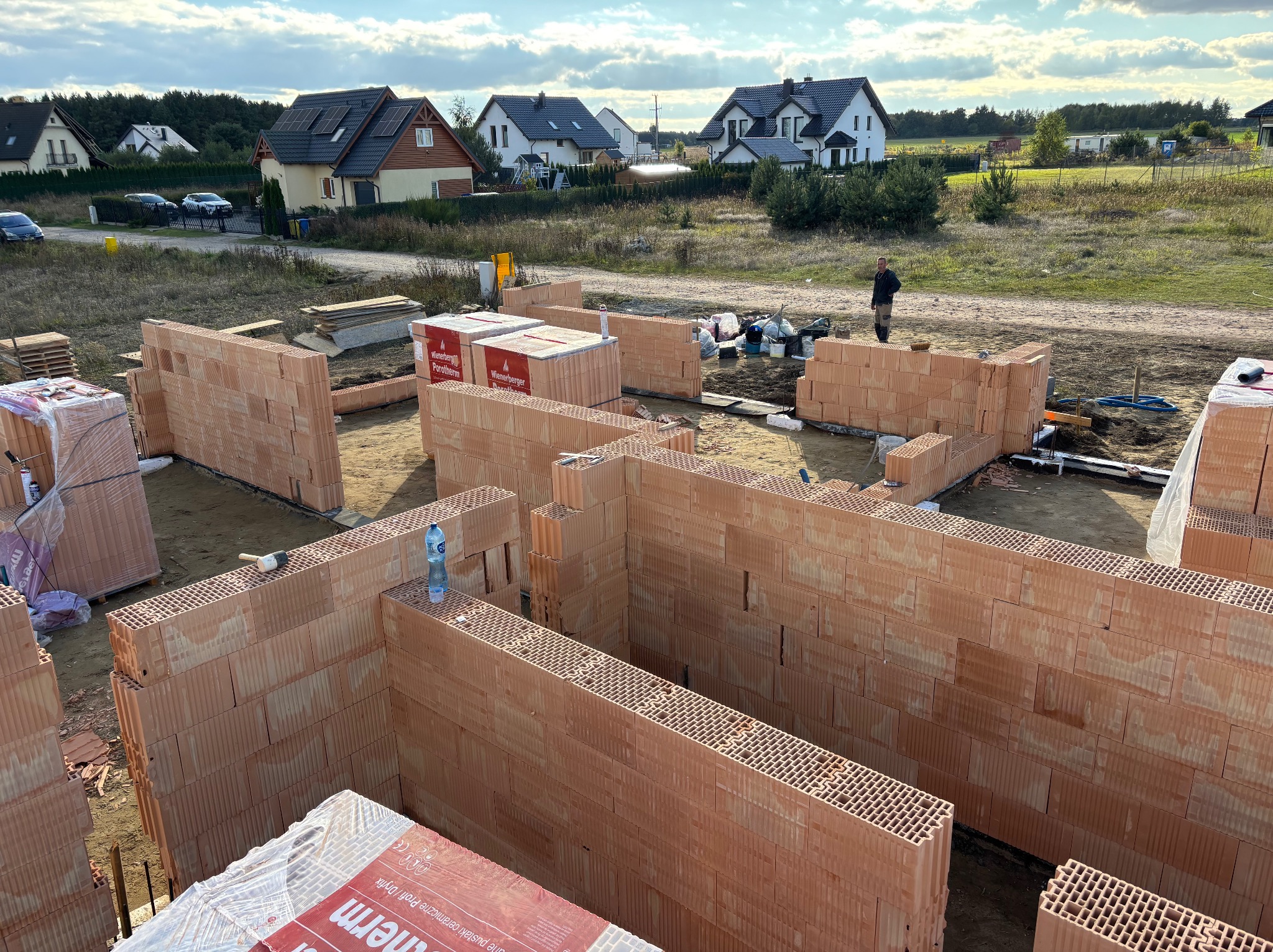 Wznoszenie ścian parteru domu z czerwonej cegły ceramicznej Porotherm. Widoczne palety z cegłami, butelka wody na murze i domy w tle.