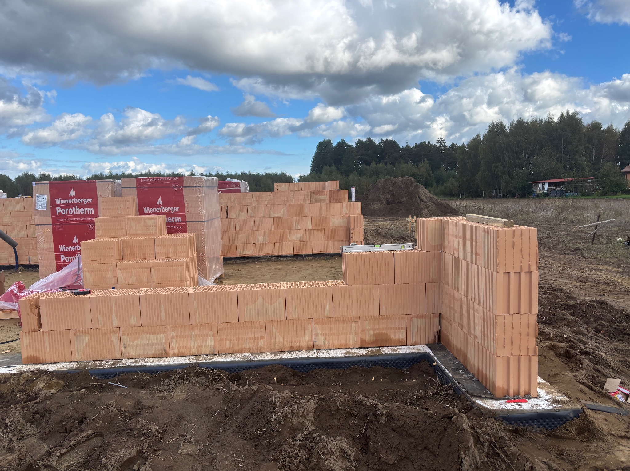 Wznoszenie ścian z pustaków Porotherm na fundamencie. Widoczne palety z materiałem budowlanym. Budowa domu w trakcie realizacji, na tle drzew i błękitnego nieba.