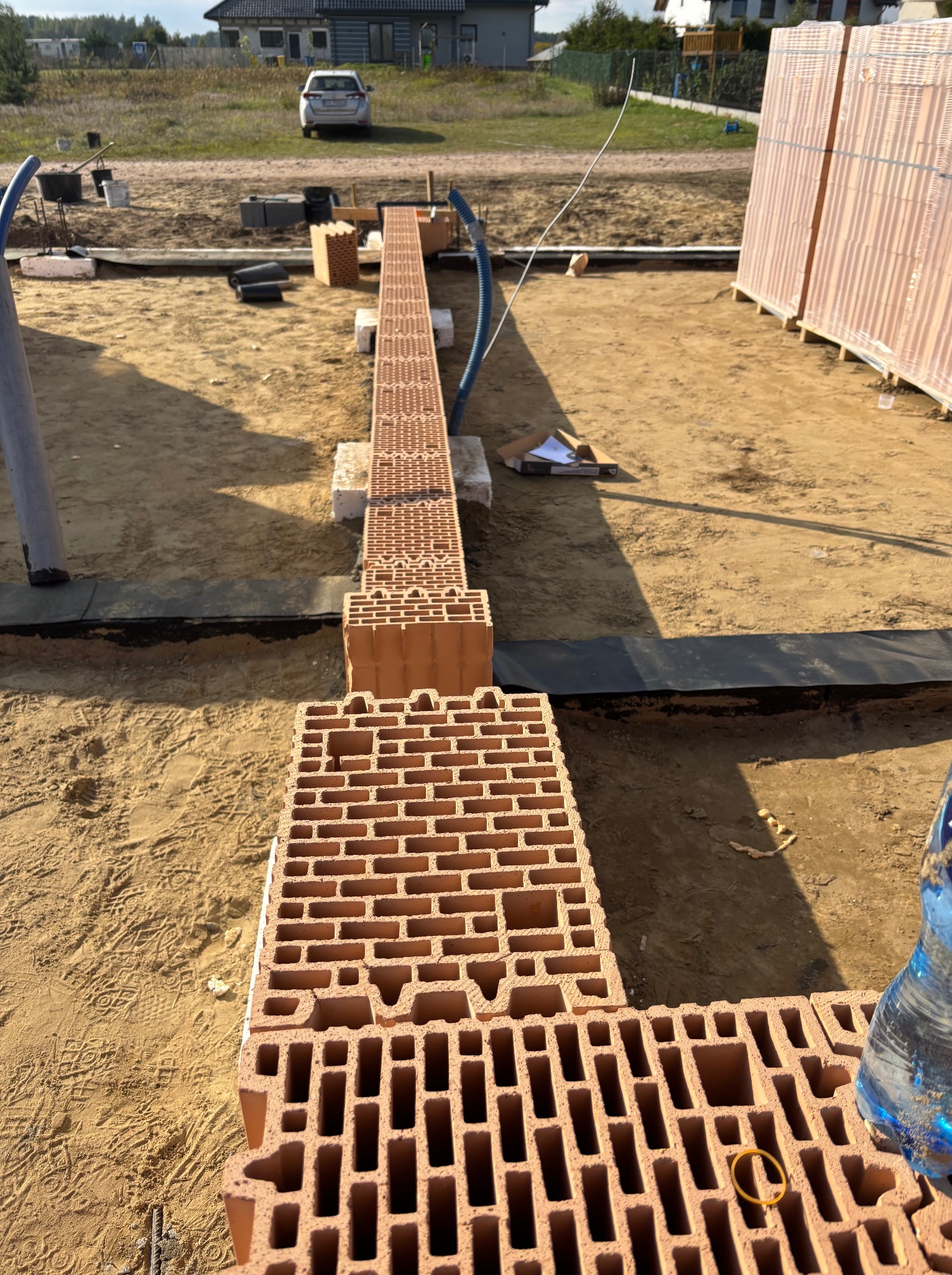 Wznoszenie fundamentów z pustaków ceramicznych na piasku, widoczne zbrojenie i izolacja. W tle dom w budowie i zaparkowany samochód.