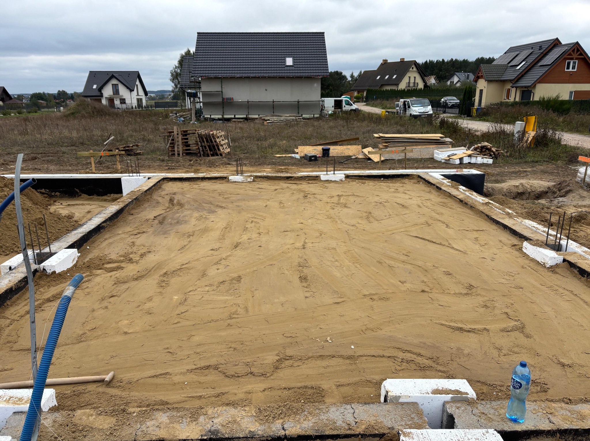 Przygotowany teren pod fundamenty domu jednorodzinnego. Widoczne zbrojenia, izolacja i piasek. W tle zabudowa jednorodzinna i pochmurne niebo.