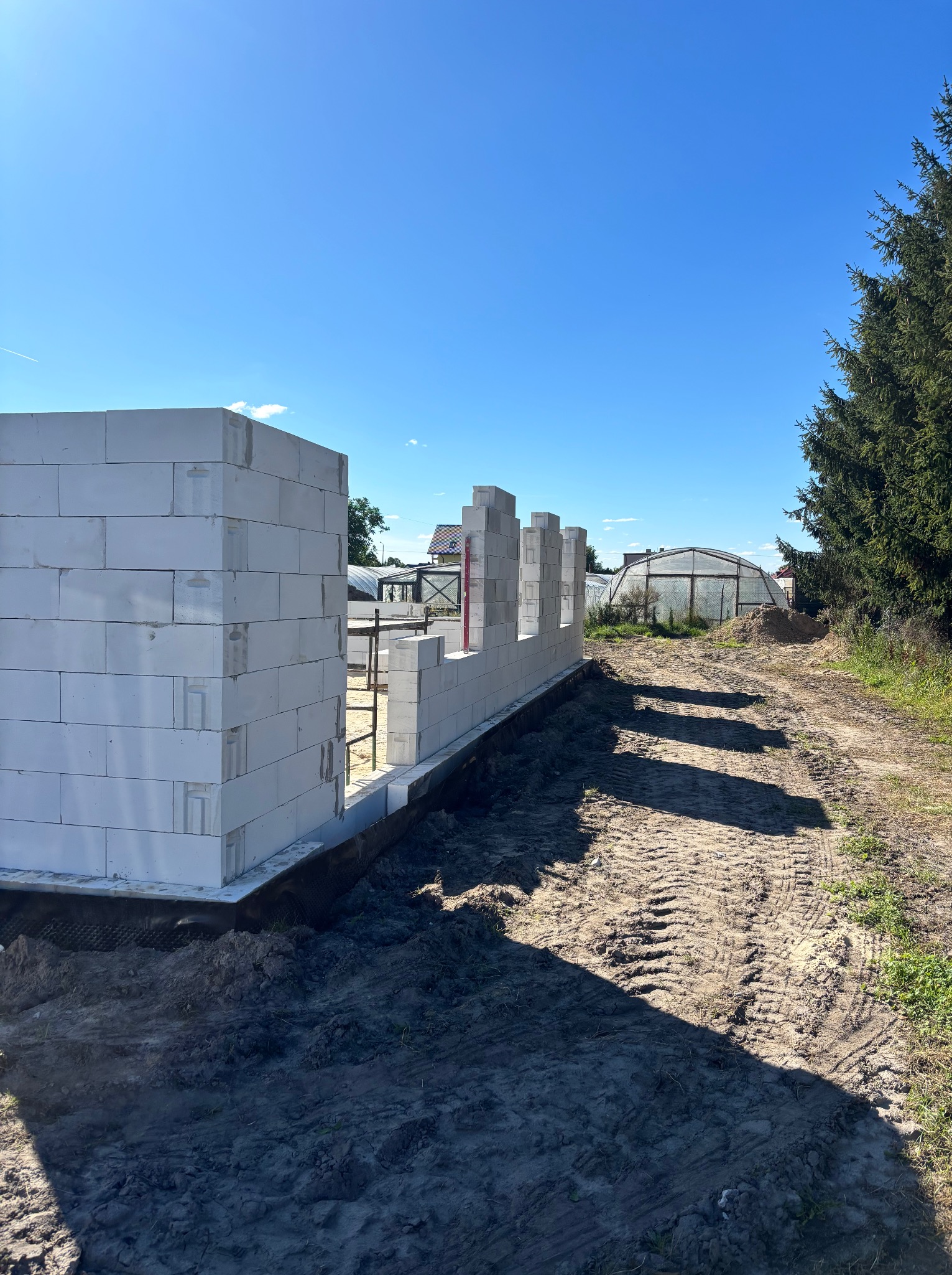 Wznoszone ściany z białych bloczków na fundamencie, widoczne z zewnątrz. W tle szklarnia i drzewa, błękitne niebo. Budowa domu w trakcie realizacji.