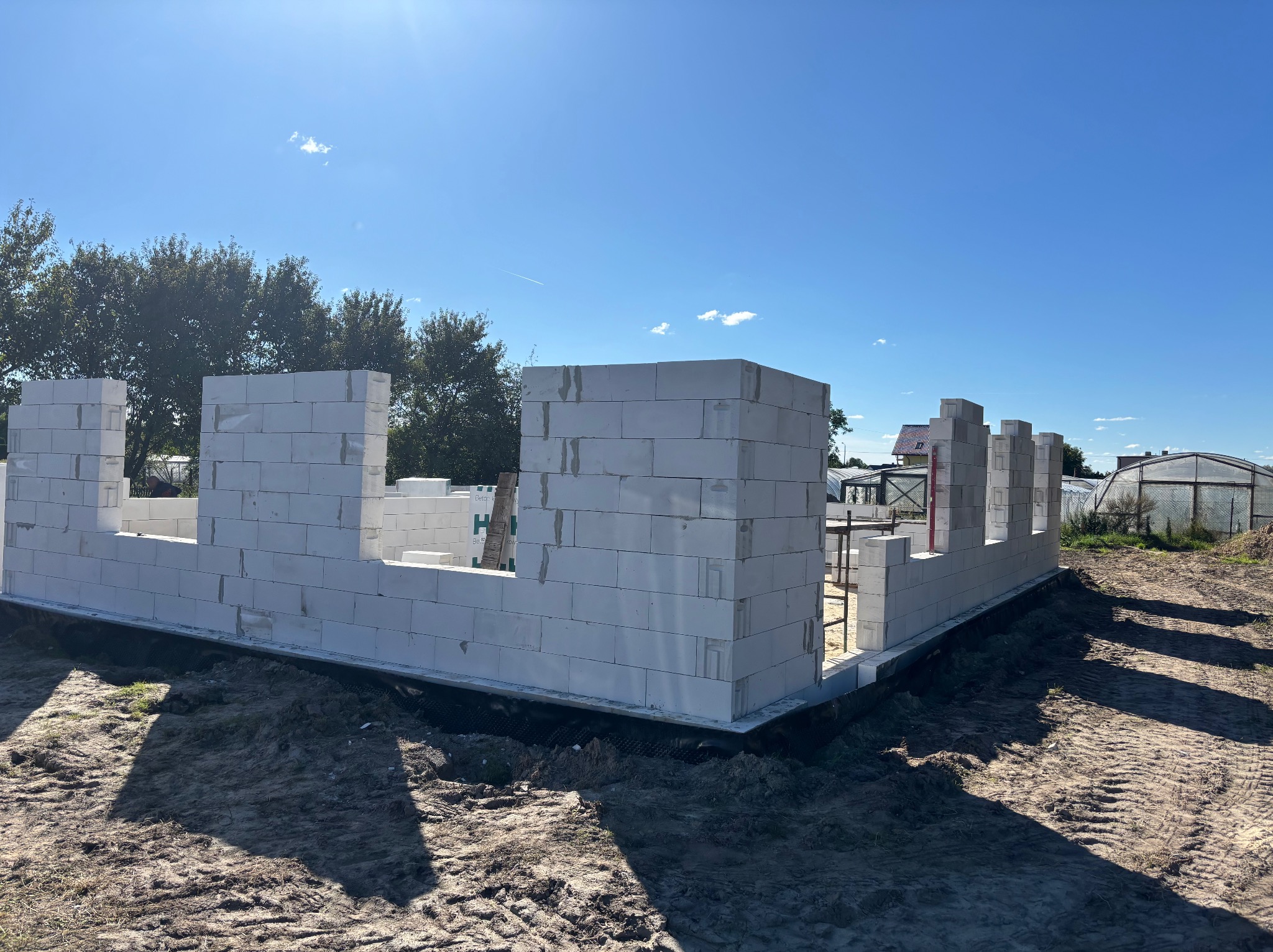 Wznoszone ściany z białych bloczków na fundamencie, widoczne zbrojenia i izolacja. W tle zieleń drzew i błękitne niebo. Budowa domu w słoneczny dzień.