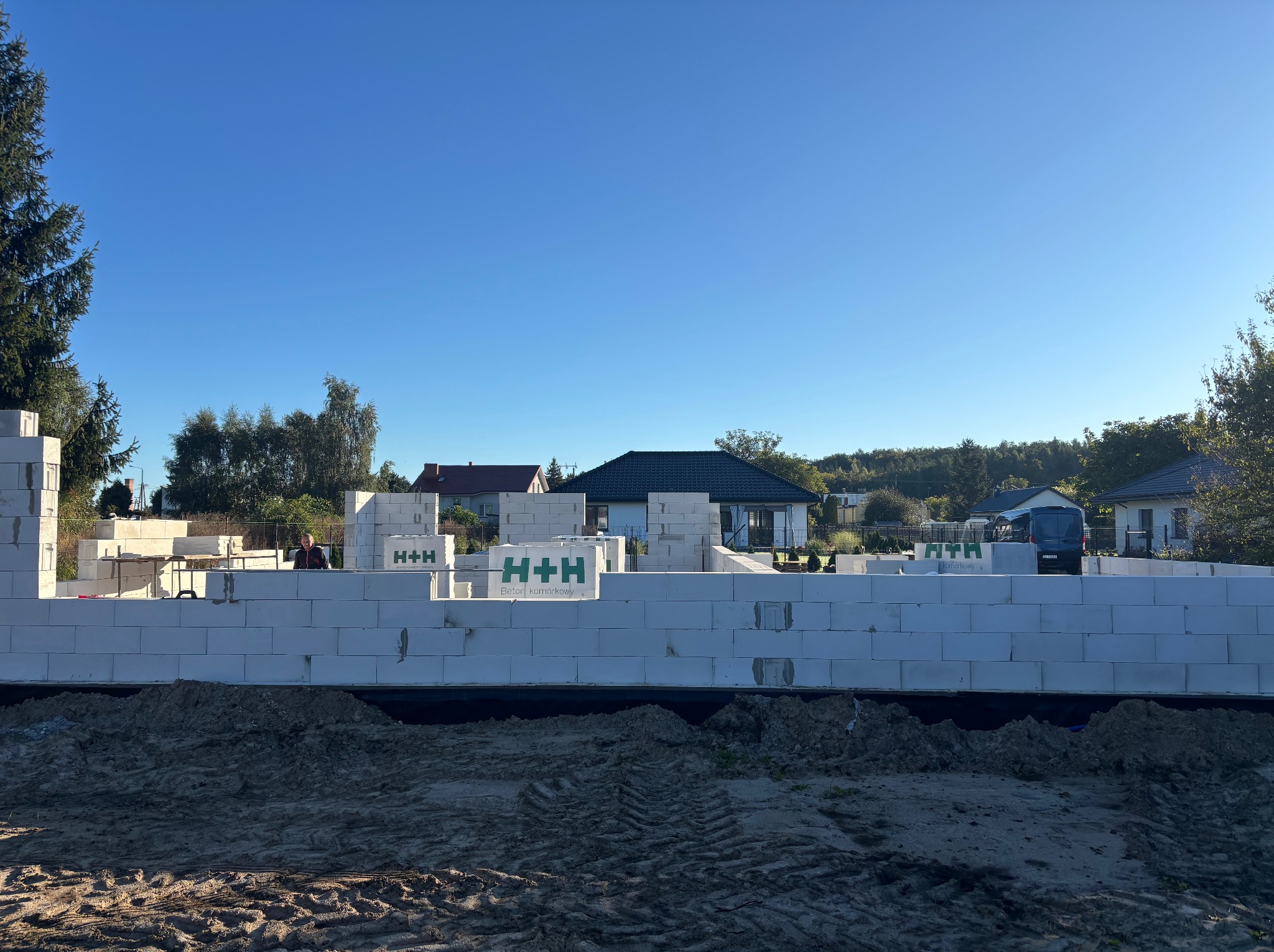 Wznoszone ściany z betonu komórkowego H+H na fundamencie. Widoczny fragment budynku w trakcie budowy, zarys murów na tle błękitnego nieba i zabudowy jednorodzinnej.