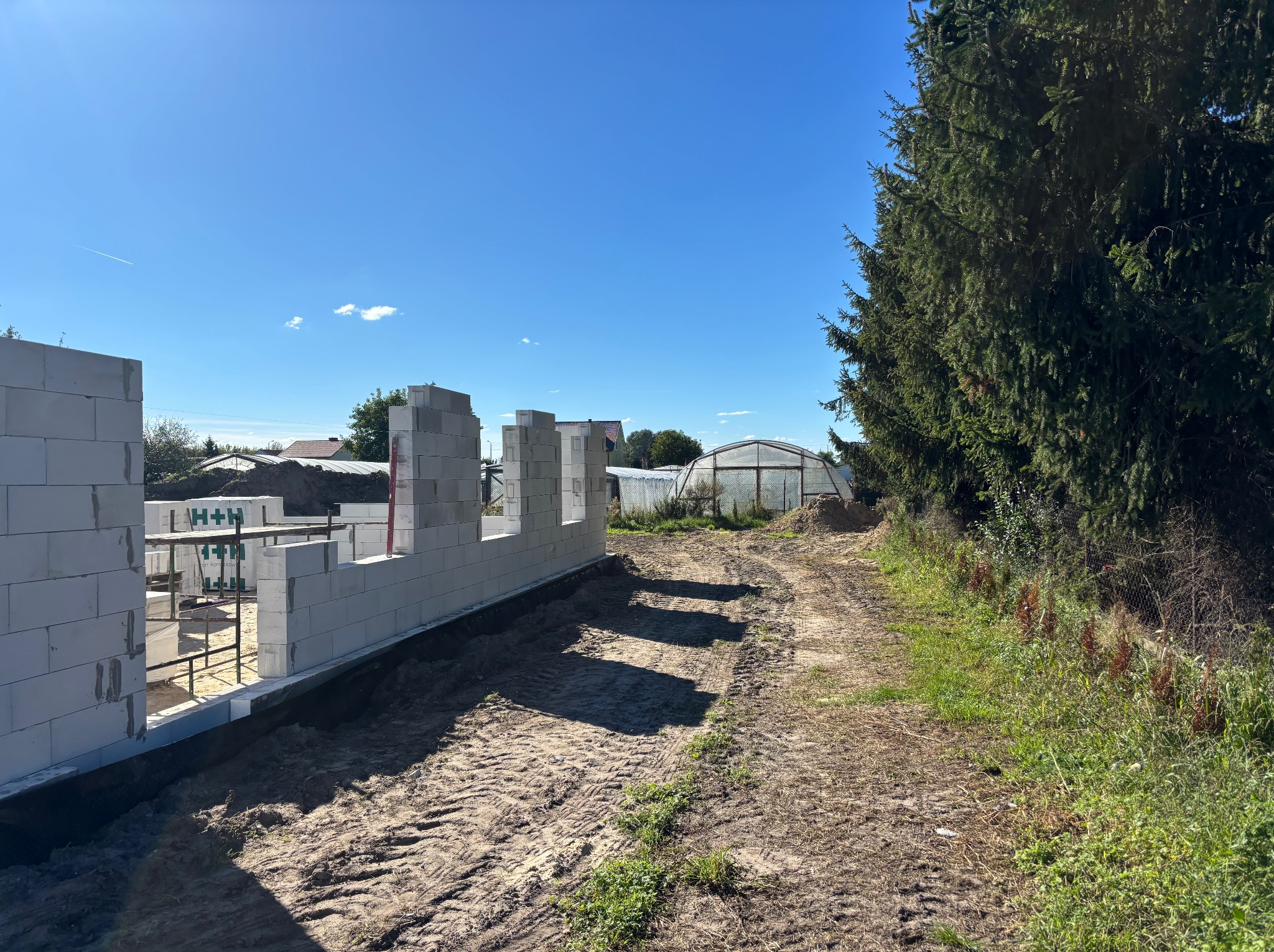 Wznoszone ściany domu z bloczków H+H na fundamencie, widok z zewnątrz. W tle szklarnia i błękitne niebo. Ślady opon na ziemi.