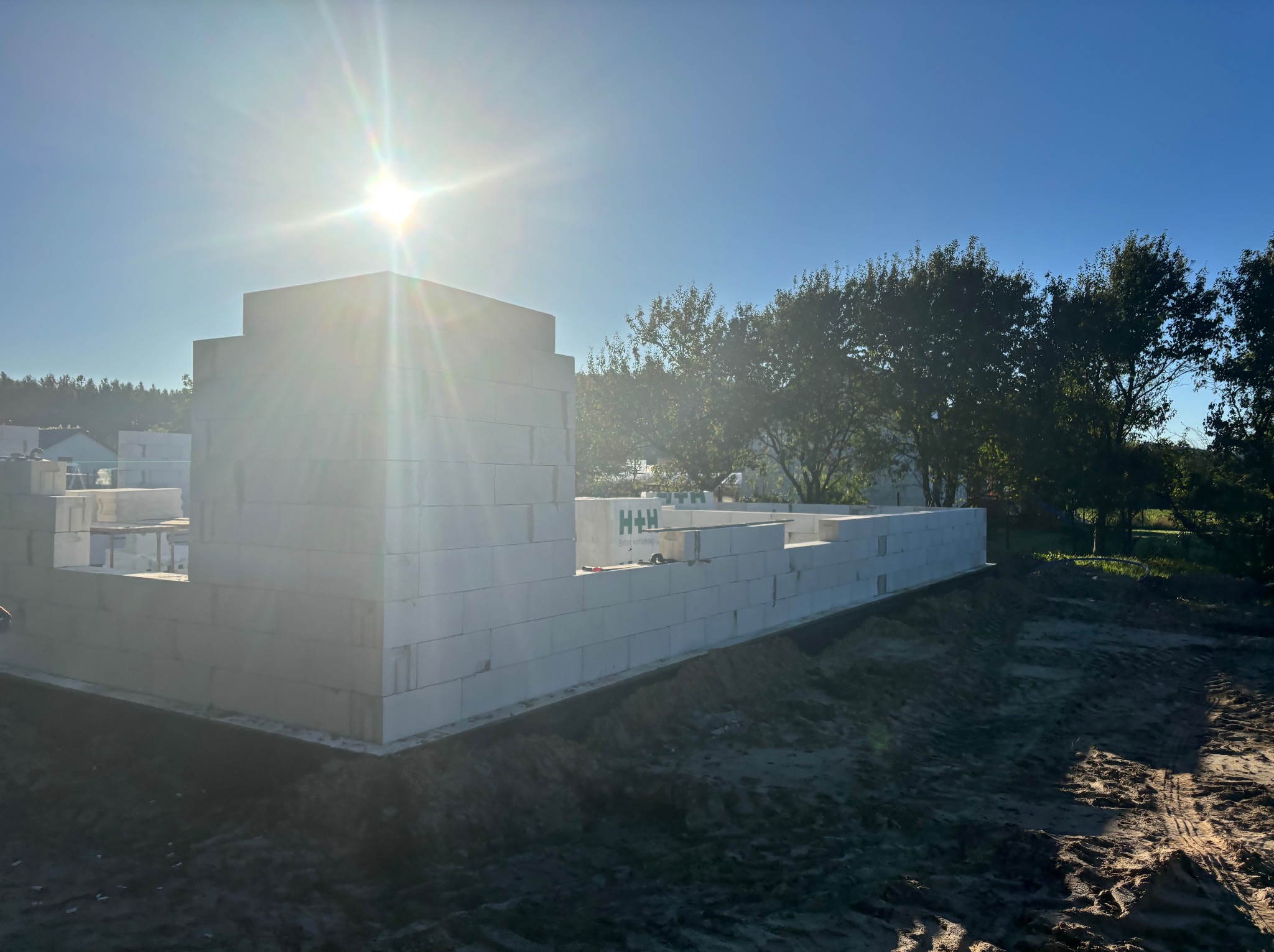 Wznoszone ściany domu z białych bloczków, widoczne narzędzia murarskie, w tle drzewa i błękitne niebo z promieniami słońca. Etap budowy w słoneczny dzień.