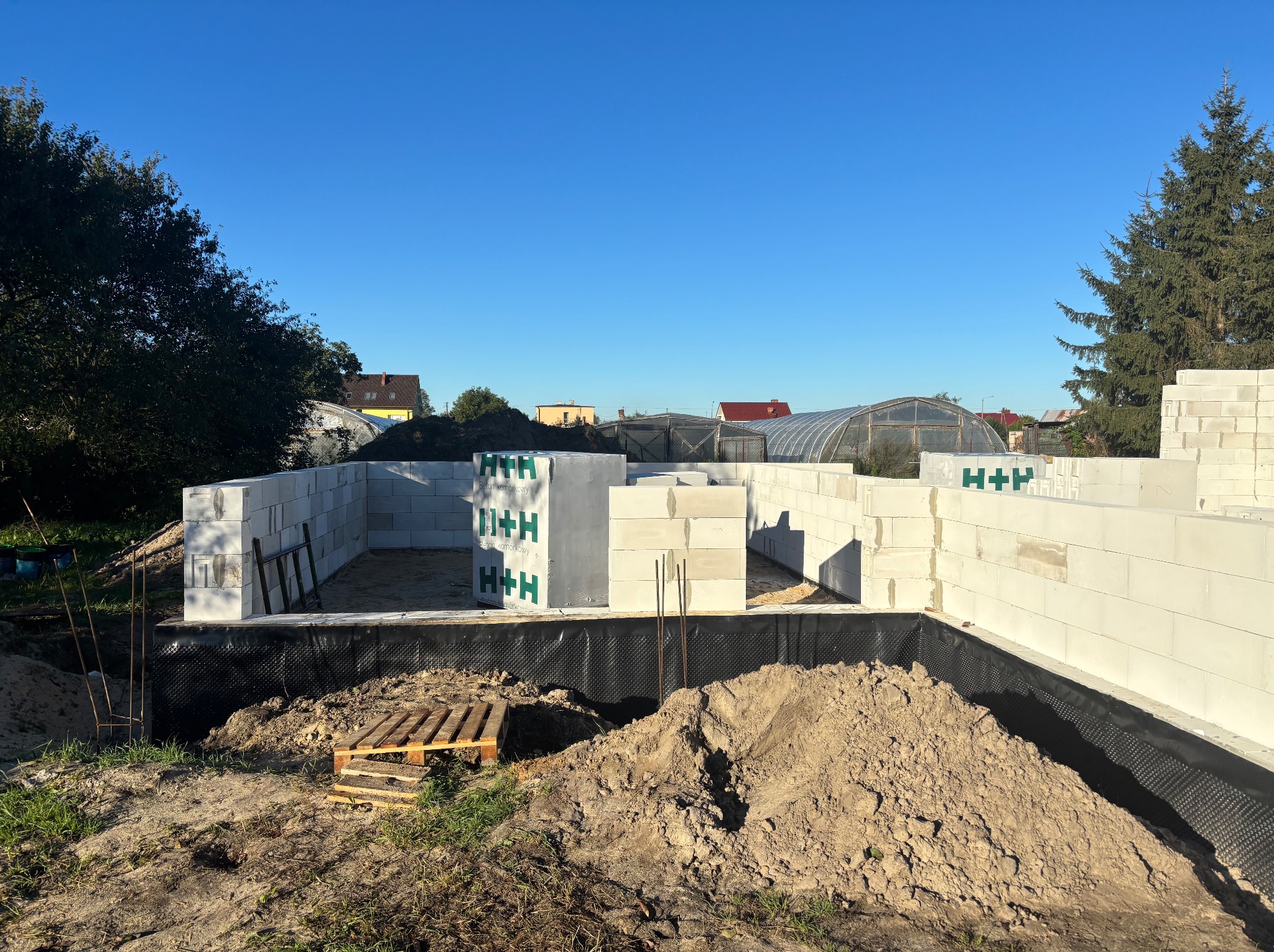 Wznoszenie ścian fundamentowych z bloczków H+H na czarnej izolacji. Widoczne sterczące pręty zbrojeniowe, paleta i pryzma ziemi. W tle zabudowa jednorodzinna i szklarnia.