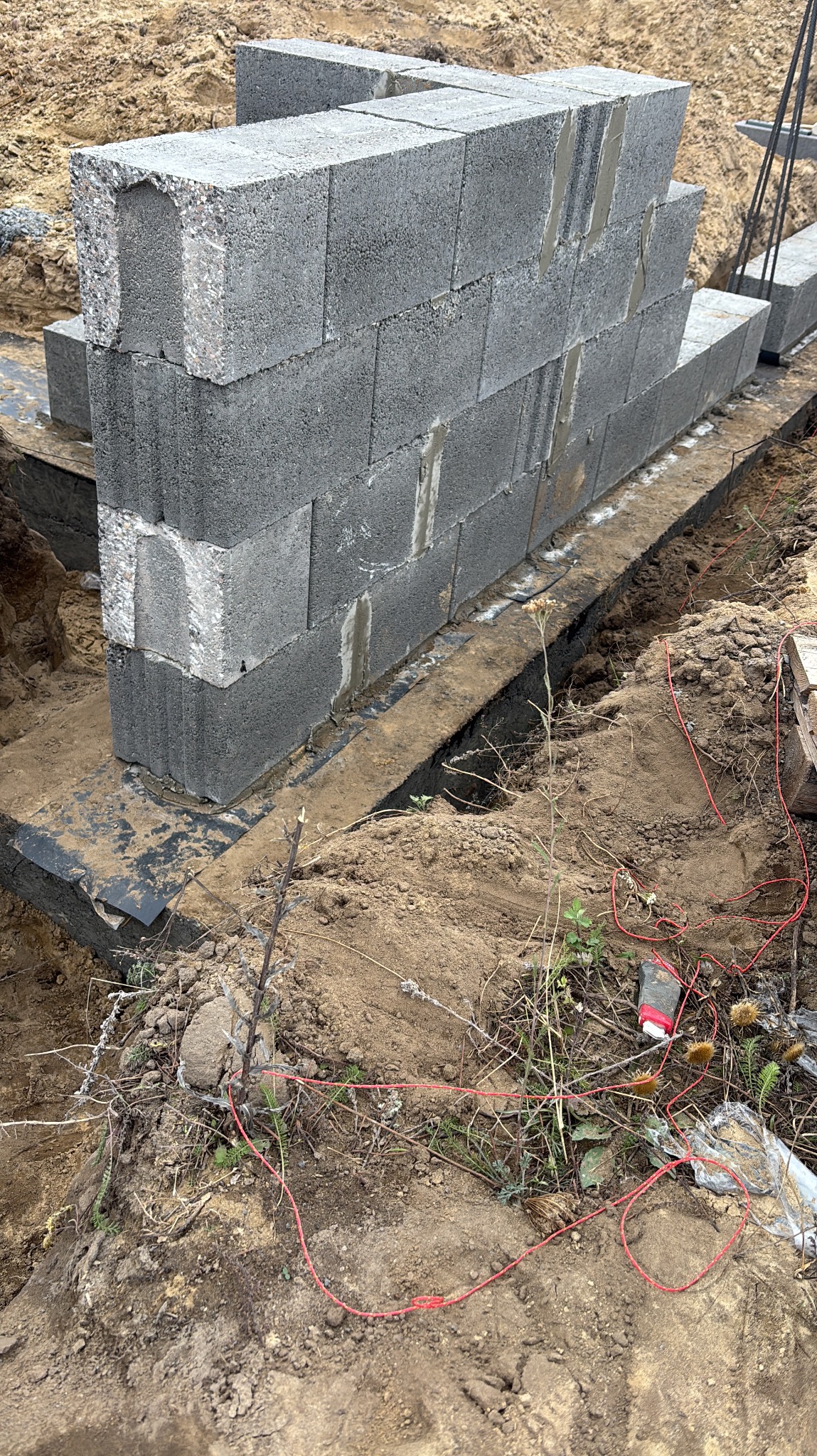 Wznoszenie ściany z szarych bloczków betonowych na fundamencie z izolacją, widoczne elementy zbrojenia i wykop, ziemia wokół.