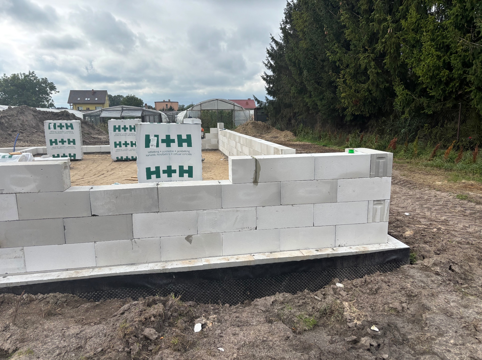 Wznoszenie ścian fundamentowych z bloczków H+H na czarnej izolacji. Częściowo wymurowane ściany, w tle palety z bloczkami i zieleń. Pochmurne niebo.