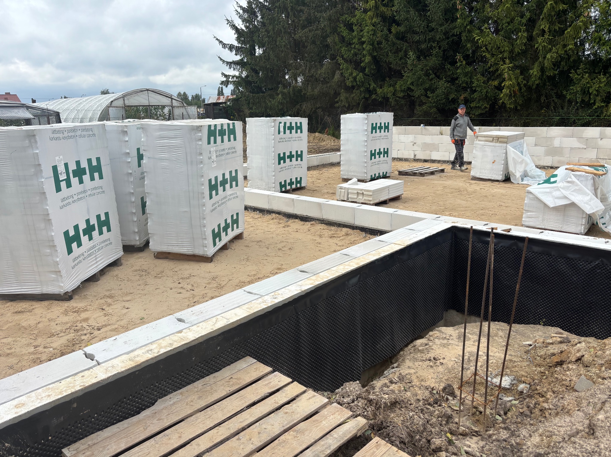 Fundamenty domu w trakcie budowy: izolacja pionowa, bloczki H+H, zbrojenie. W tle pracownik budowlany i szklarnia. Ujęcie z bliska, widoczne detale izolacji.
