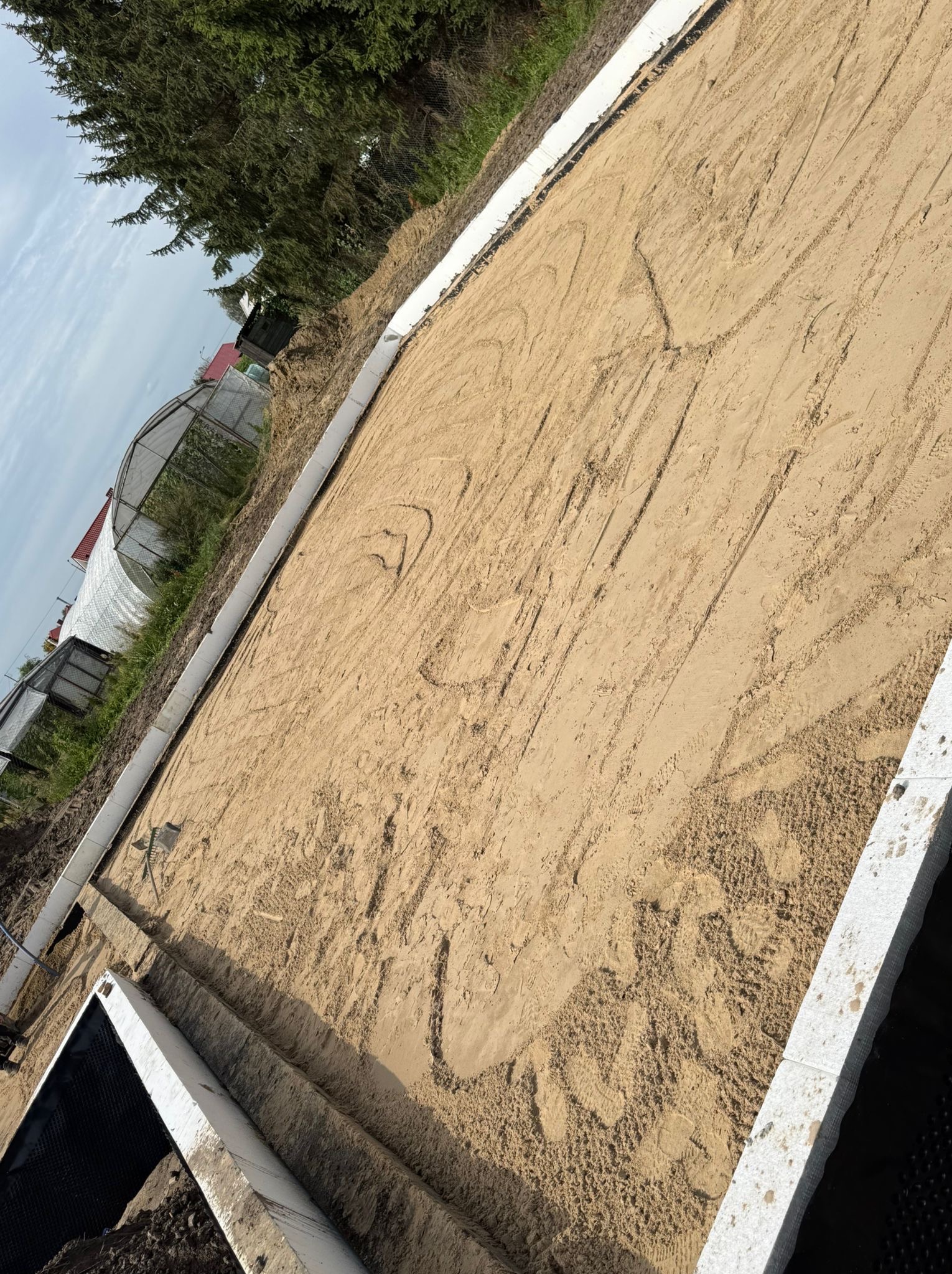 Przygotowany teren pod fundament z widocznymi obrysami i piaskiem, otoczony białym obramowaniem. W tle widoczny tunel foliowy i zieleń.