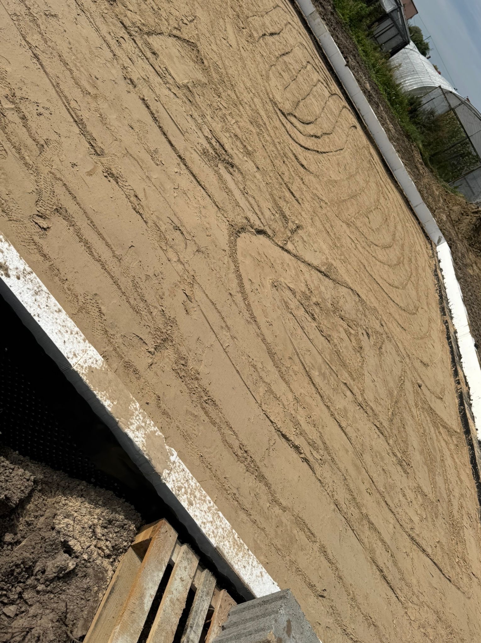 Przygotowany teren pod fundamenty z widoczną izolacją pionową i obrzeżami ze styropianu. Ślady opon na piasku. W tle zabudowania.