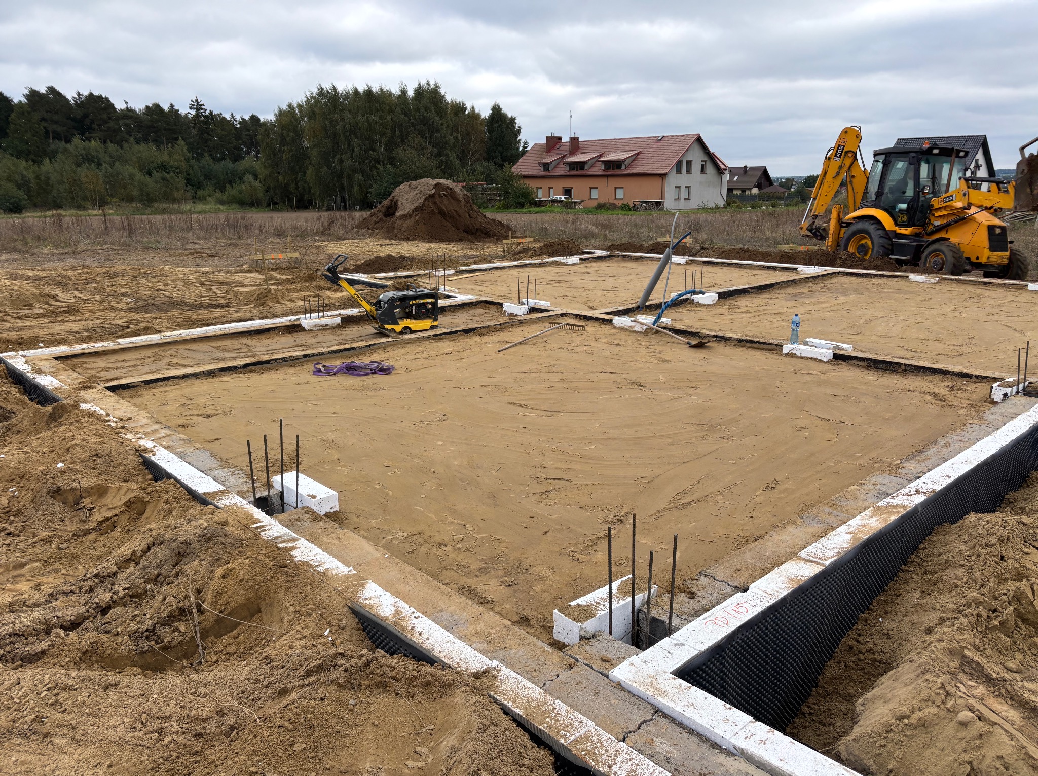 Zarys fundamentów domu z widoczną izolacją pionową, zbrojeniami i sprzętem budowlanym w tle. Ujęcie z góry, w tle dom i drzewa.