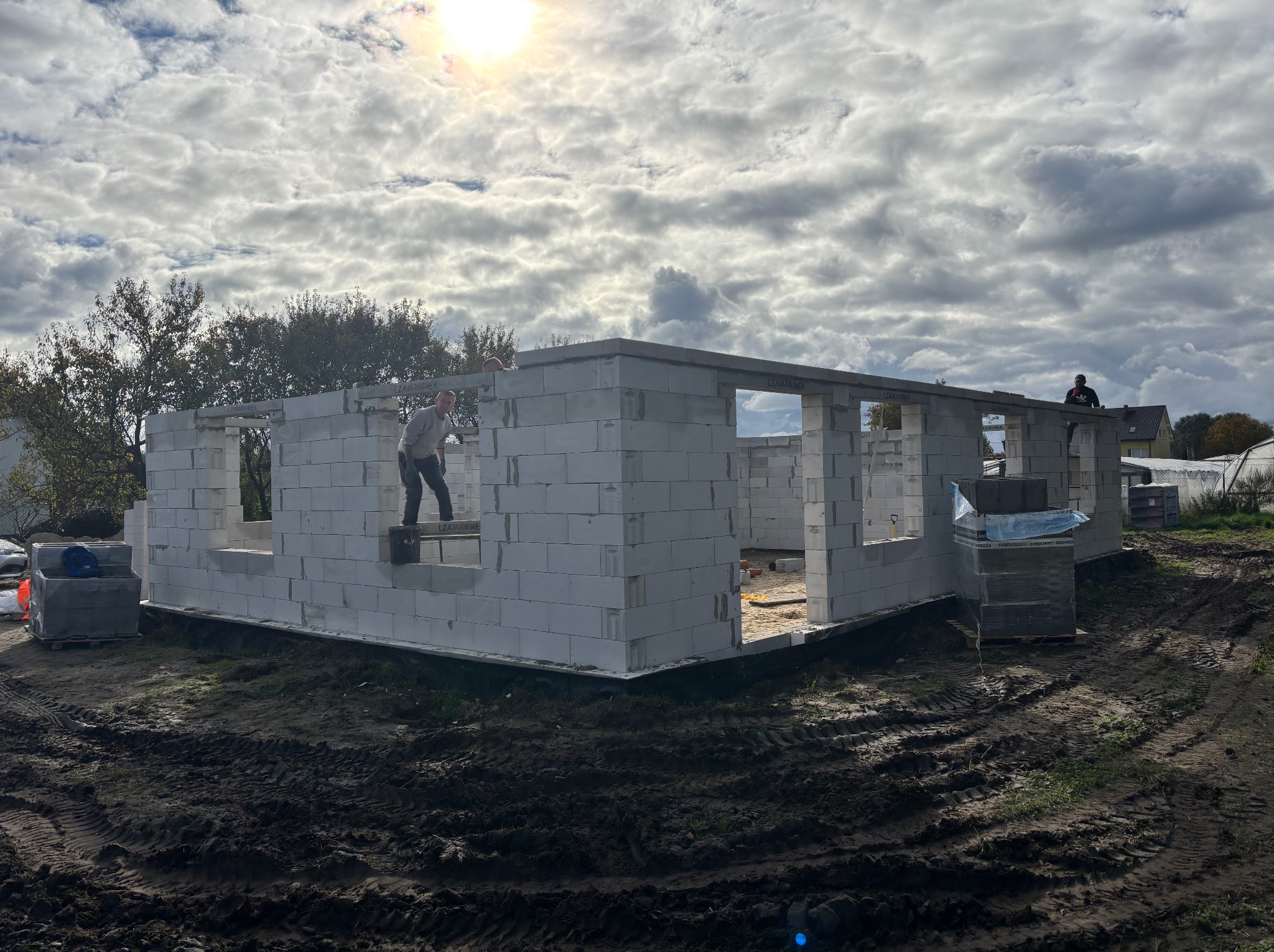 Wznoszenie ścian parterowego domu z betonu komórkowego. Dwóch pracowników na budowie, palety z bloczkami. Widok na fundamenty i częściowo postawione ściany.