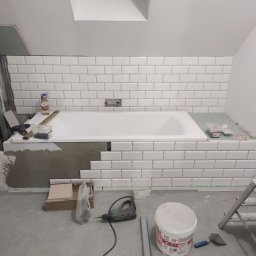 Alexstar Bud Company Oleksandr Nahliuk - Łazienka w trakcie remontu: białe, prostokątne płytki ułożone nad wanną, narzędzia glazurnicze widoczne na podłodze i wannie. Widoczne pionowe poziomice.