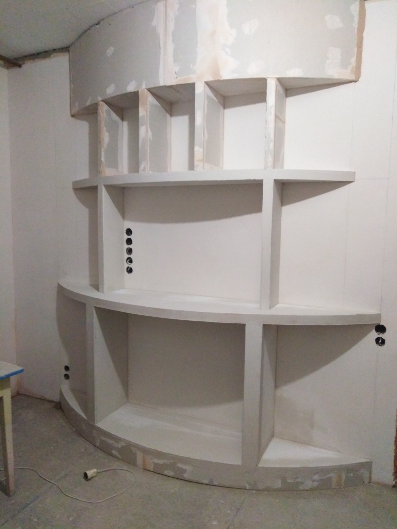 Narożna zabudowa z płyt gipsowo-kartonowych w trakcie montażu, z widocznymi otworami na gniazdka elektryczne i niedoskonałościami wykończenia. Surowy stan wnętrza.