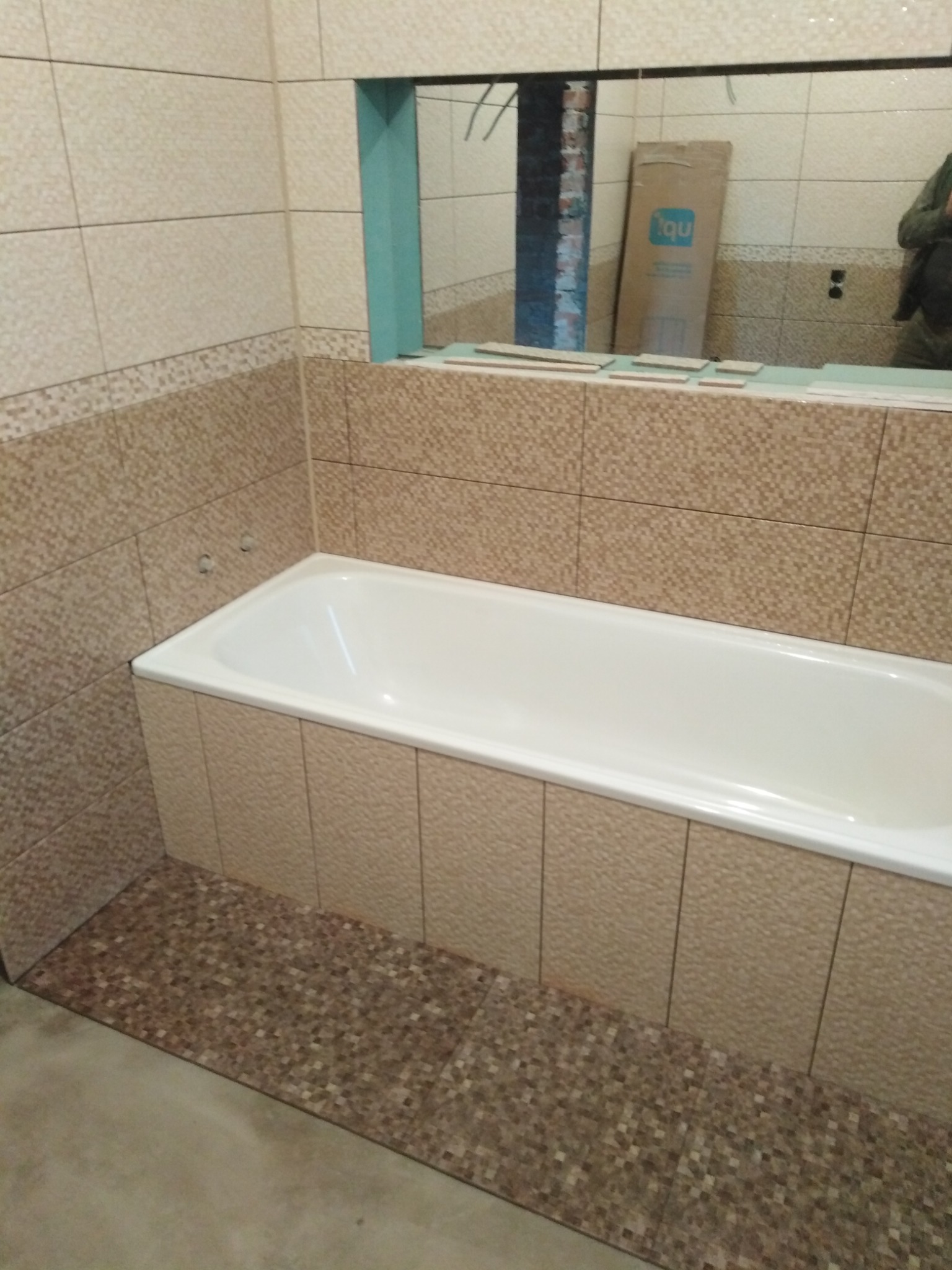 Jasna łazienka w trakcie wykańczania: beżowe płytki na ścianach i mozaika na podłodze, biała wanna obudowana płytkami. Widoczne przygotowania do montażu lustra.