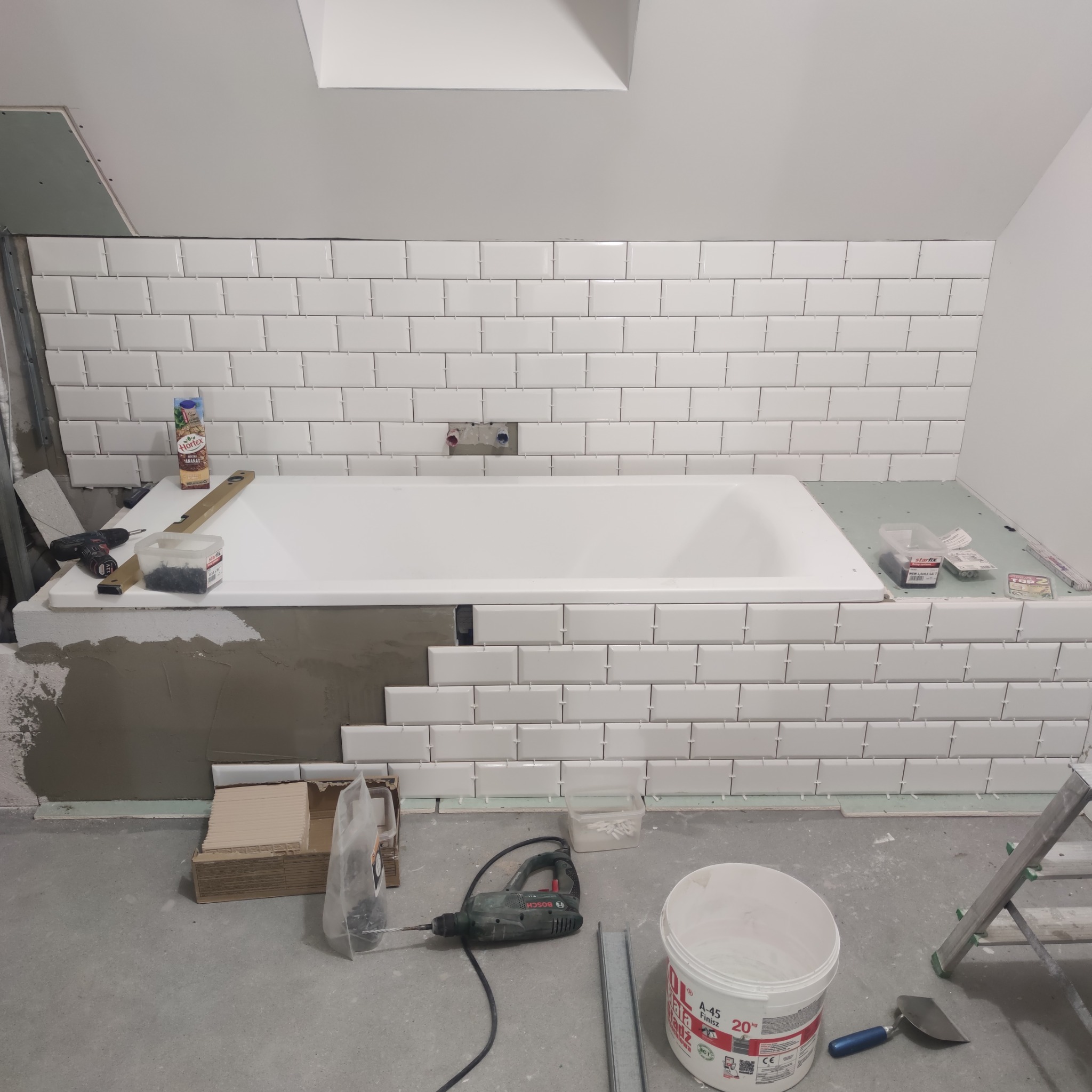 Łazienka w trakcie remontu: białe, prostokątne płytki ułożone nad wanną, narzędzia glazurnicze widoczne na podłodze i wannie. Widoczne pionowe poziomice.
