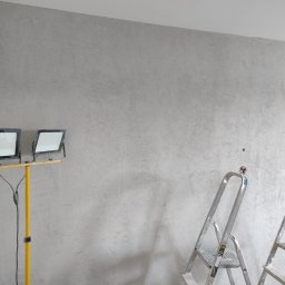 Frontmen - Ściana po malowaniu dekoracyjnym w odcieniach szarości, oświetlona lampami budowlanymi, z drabinami w tle. Efekt wykończenia wnętrza.