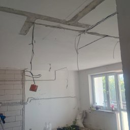Frontmen - Surowy stan wnętrza w trakcie remontu: widoczne przewody elektryczne na ścianach i suficie, niedokończone ściany z pustaków oraz okno z widokiem na zieleń.