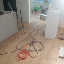 Frontmen - Montaż szafki na drewnianej podłodze, w tle pracownik przycina element. Widoczne kable, narzędzia i akcesoria montażowe. Wnętrze w trakcie wykańczania.