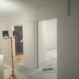 Frontmen - Białe wnętrze w trakcie malowania z lampą na statywie, widoczne zabezpieczenia podłogi folią. W tle otwarte przejście do kolejnego pomieszczenia.