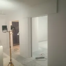 Białe wnętrze w trakcie malowania z lampą na statywie, widoczne zabezpieczenia podłogi folią. W tle otwarte przejście do kolejnego pomieszczenia.