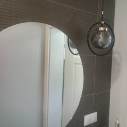 Frontmen - Nowoczesna łazienka z okrągłym lustrem, szarą, teksturowaną ścianą z płytek i designerską lampą wiszącą. Widoczne gniazdka i włącznik. Minimalistyczny styl.