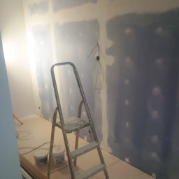 Frontmen - Pokój w trakcie szpachlowania ścian, widoczna drabina malarska, wiaderka z masą szpachlową. Ściany częściowo pokryte szpachlą, widoczne punkty elektryczne.