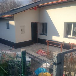 Frontmen - Elewacja domu po malowaniu z widocznymi puszkami farby i rusztowaniem. Świeżo pomalowana dolna część budynku na czarno. Widok zza ogrodzenia.