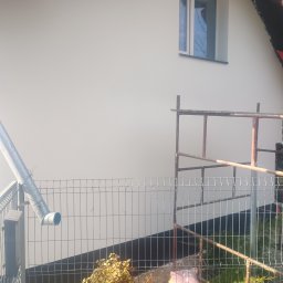 Frontmen - Świeżo pomalowana, biała ściana domu z oknem, widoczna zza metalowego ogrodzenia. Obok ściany stoi rusztowanie. Rynna i fragment dachu w kadrze.