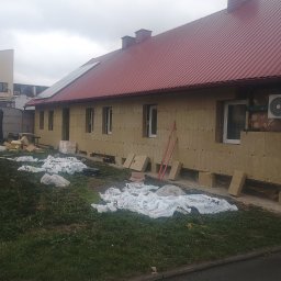 Frontmen - Budynek z czerwoną blachodachówką w trakcie ocieplania wełną mineralną. Widoczne panele słoneczne na dachu i materiały budowlane na trawniku. Prace izolacyjne w toku.
