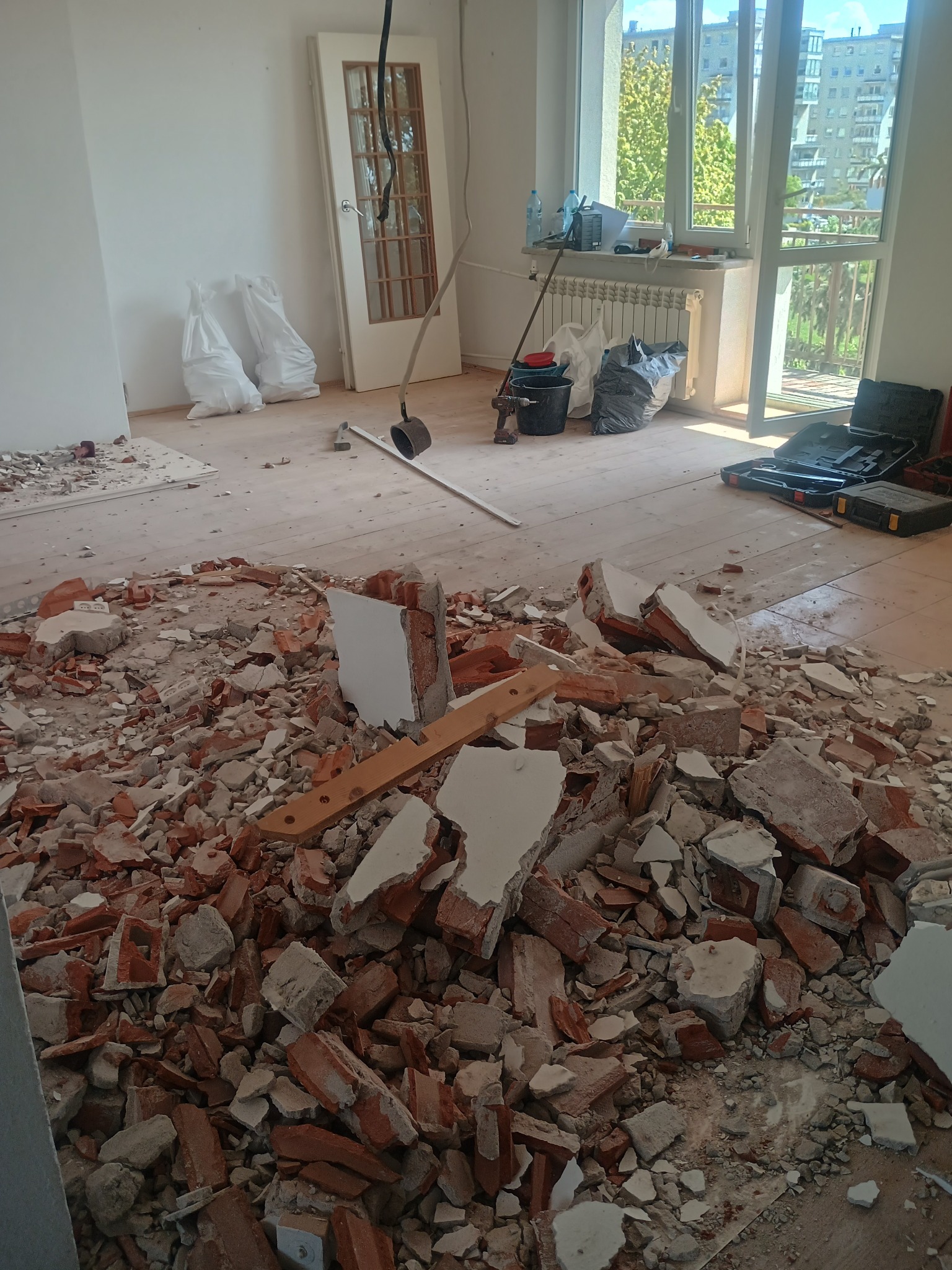 Gruz po wyburzeniach na podłodze z desek w jasnym pokoju. Widoczne okno, drzwi i narzędzia. Etap prac remontowych w mieszkaniu.