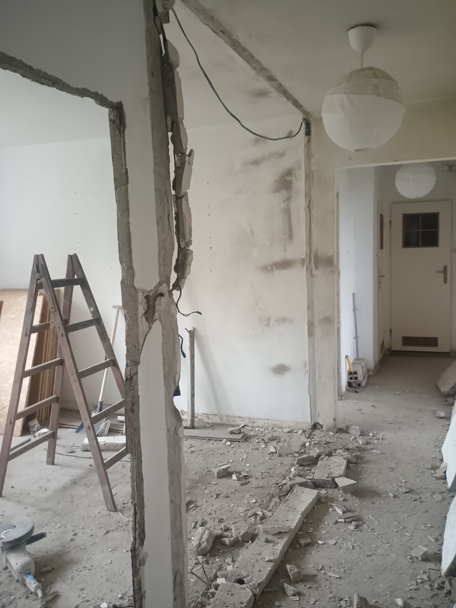 Demontaż ściany działowej w mieszkaniu. Widoczne gruz, drabina i narzędzia. Prace w toku, odsłonięte przewody elektryczne, surowy stan.