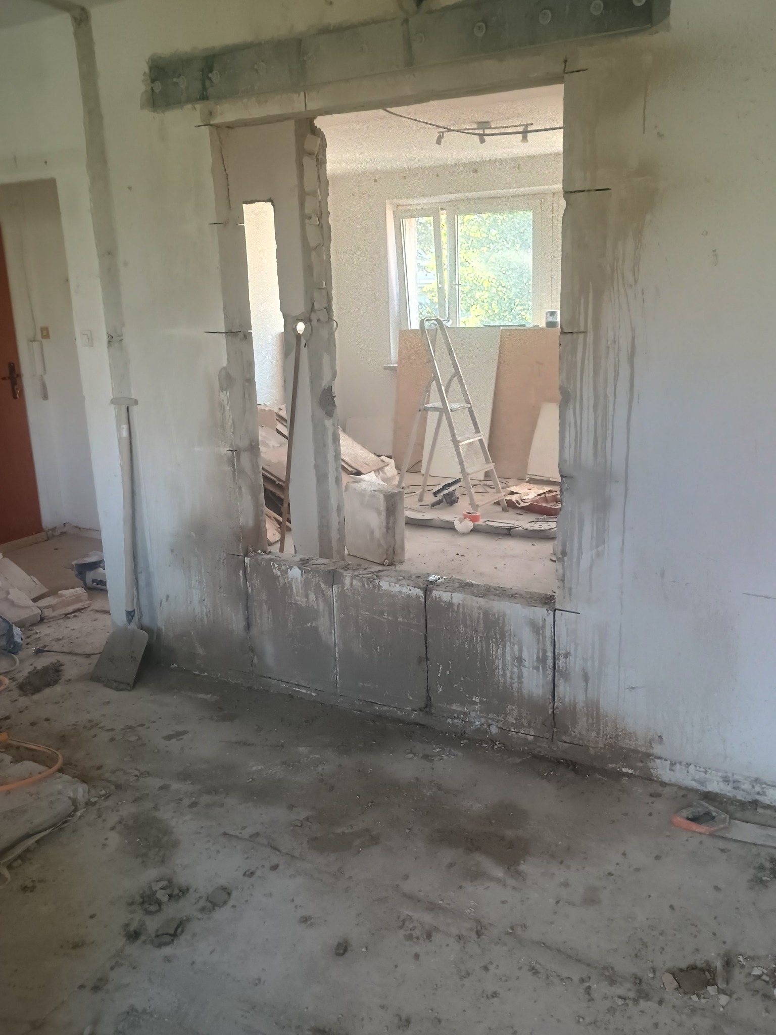 Częściowo wyburzona ściana działowa z widocznym nadprożem, gruz i narzędzia na podłodze. Widok na sąsiednie pomieszczenie z oknem i drabiną.